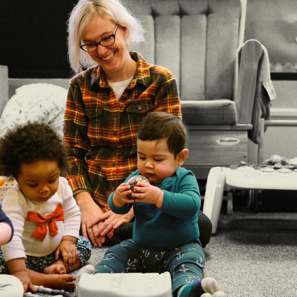 Woman smiling as she watches two children playing. They are in full color, the background is black and white.